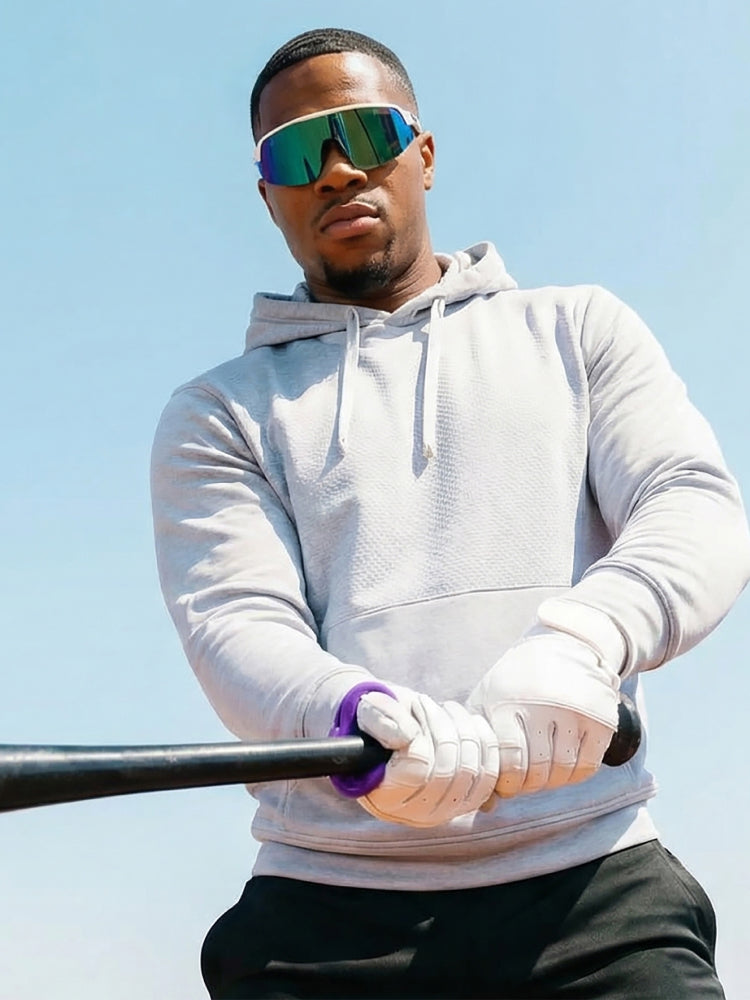 Person wearing a light gray hoodie and sunglasses, holding a baseball bat, wearing a purple hit hero against a clear blue sky.