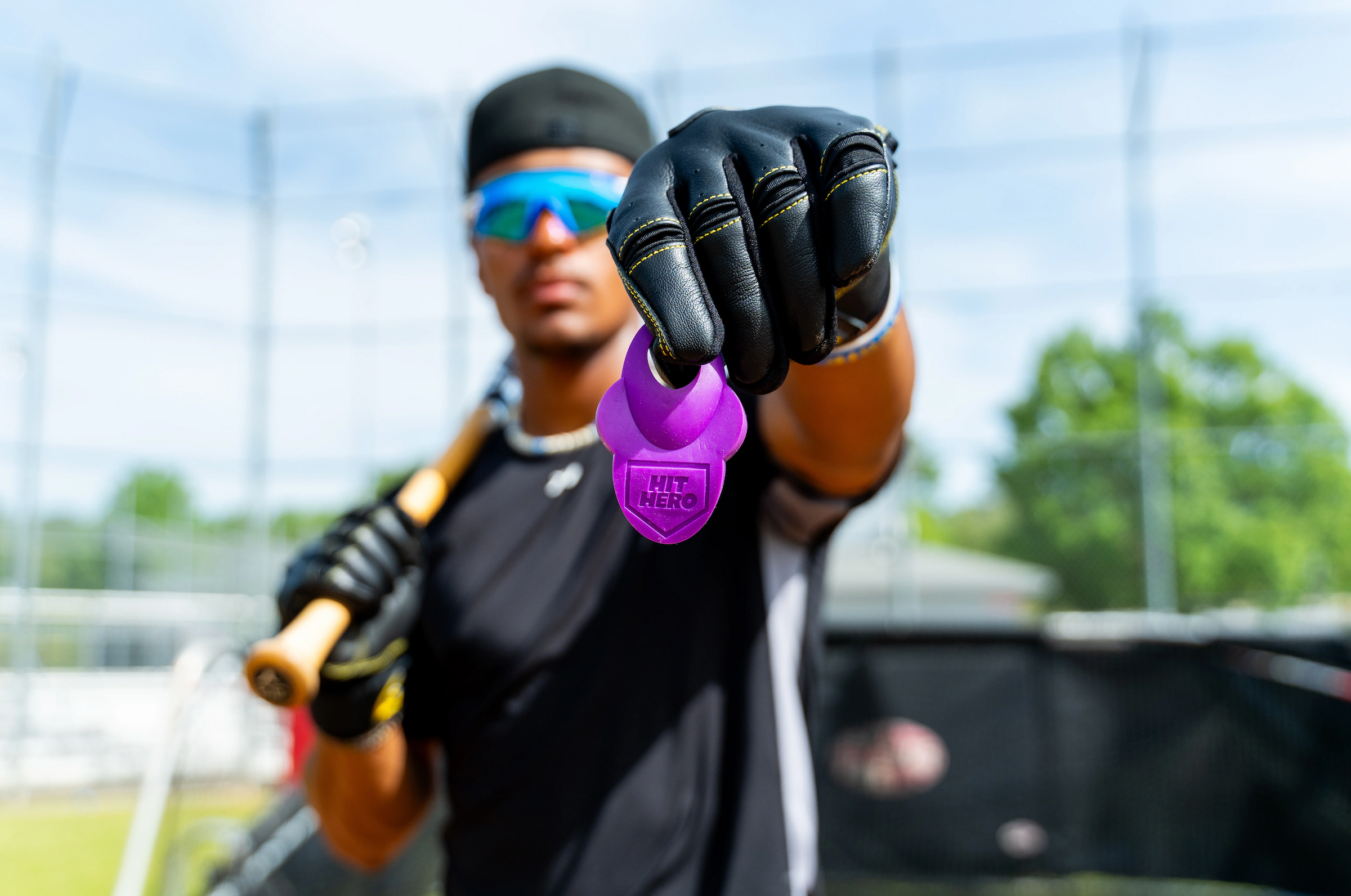 A close-up lifestyle shot of a baseball player wearing a black beanie, blue reflective sunglasses, and a black baseball glove, holding a purple 'HIT THE BALL' weight in front of his face. He is holding a wooden bat behind his shoulder, set against a blurred outdoor baseball field background.