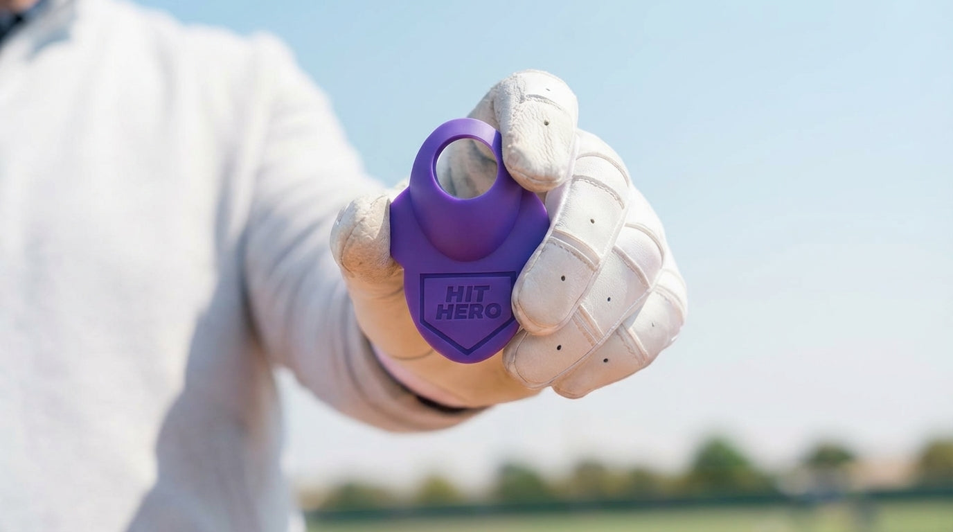 Hand holding a purple device labeled 'HIT HERO' baseball thumb guard against a blurred outdoor background
