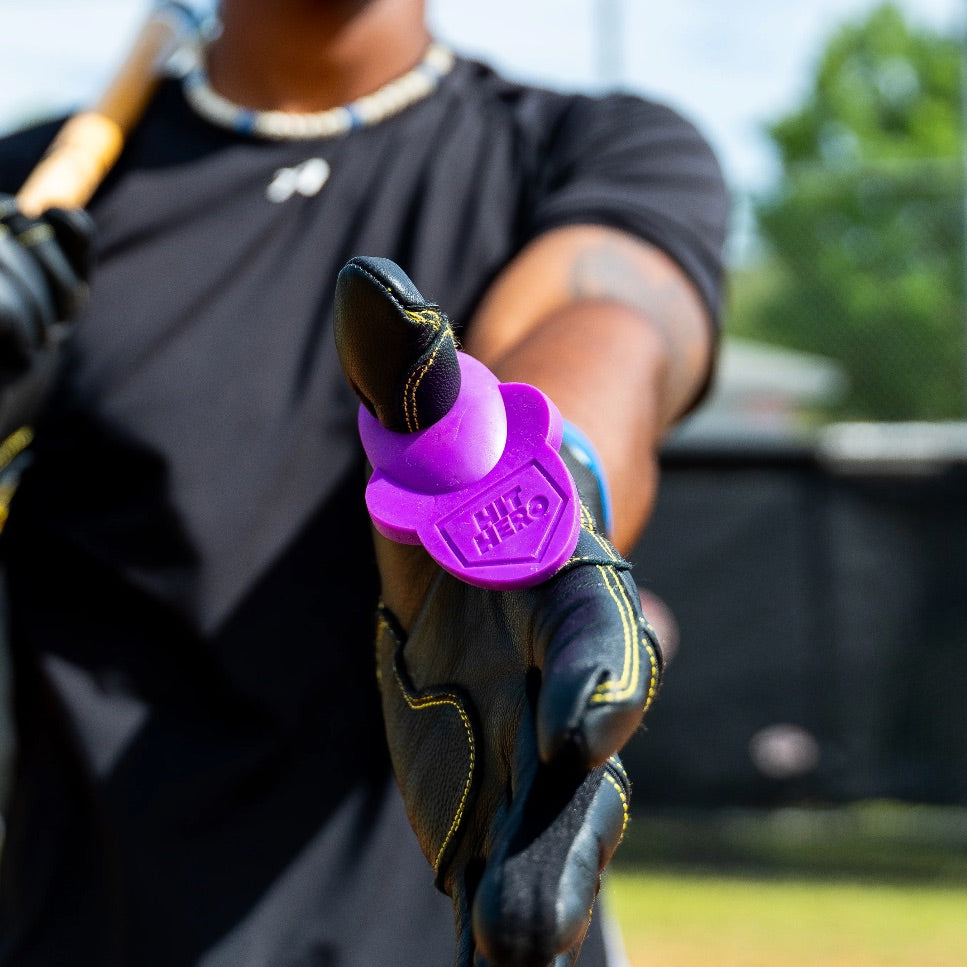 A magenta Hit Hero worn on the thumb of a black batting glove, hand held out toward the camera